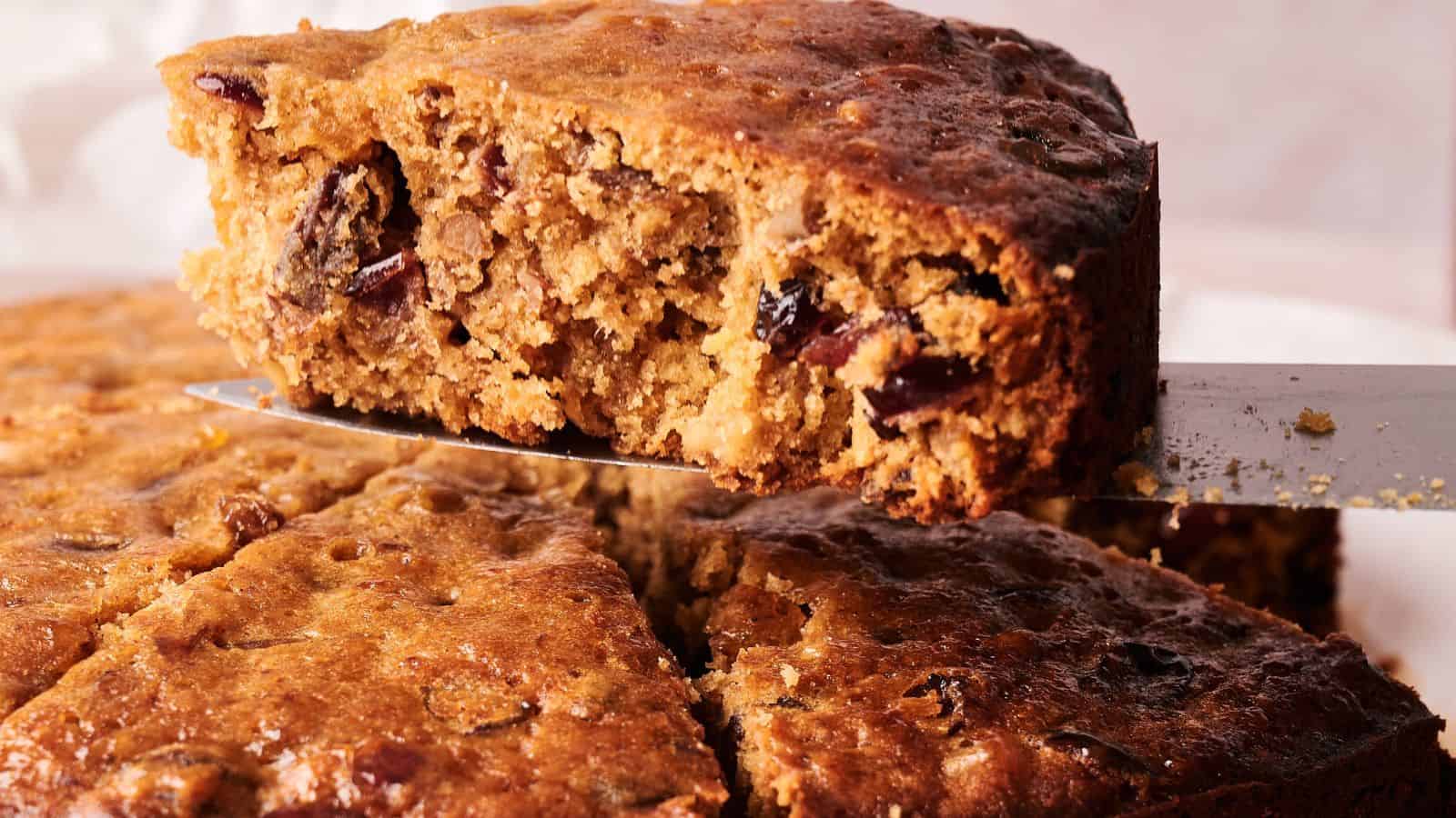 A close-up of a fruitcake with a slice being lifted out, showing its dense texture and pieces of dried fruit inside.