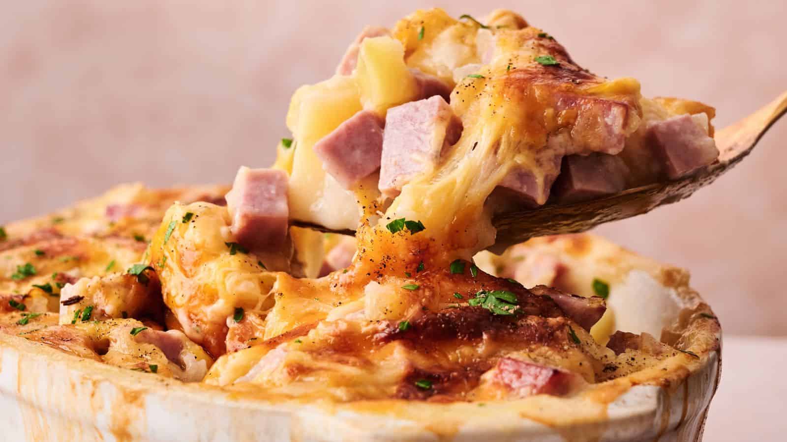 A wooden spoon lifts a serving of cheesy casserole with chunks of ham and potatoes from a baking dish.
