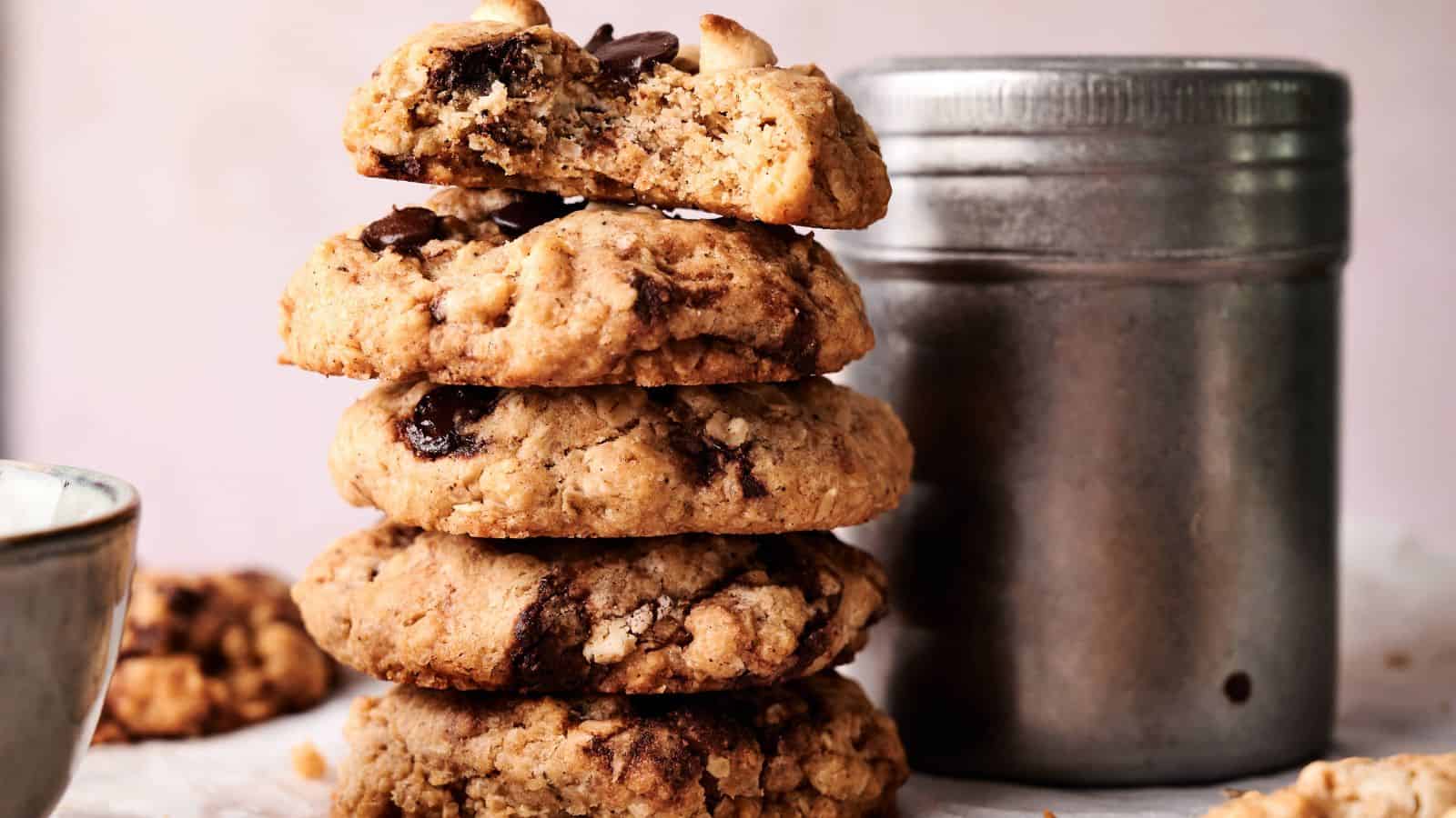 A stack of four chocolate chip cookies, with the top cookie partly broken, next to a metal container and a ceramic bowl on a light surface.