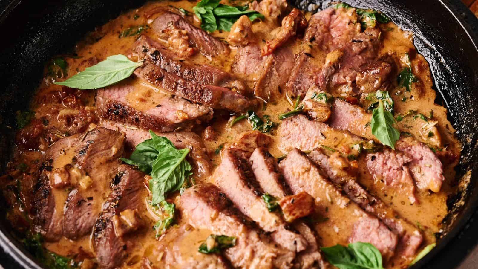 Slices of steak arranged in a creamy sauce with herbs and garnished with fresh basil leaves in a black skillet.