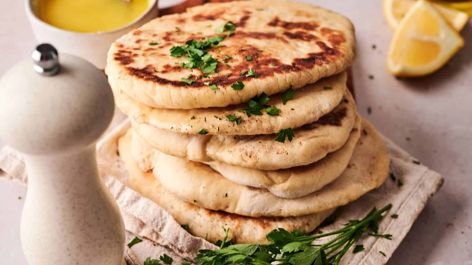 A stack of flatbreads garnished with chopped parsley sits on a cloth, with a white pepper grinder, fresh herbs, a lemon wedge, and a bowl of yellow sauce nearby.