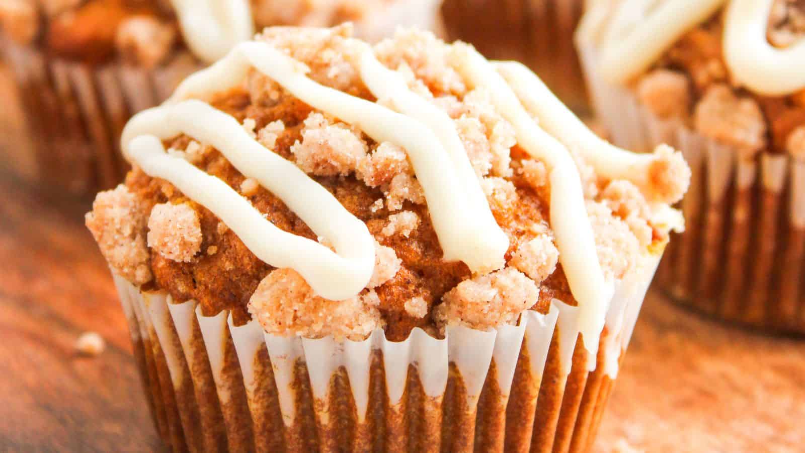 Close-up of a muffin topped with crumbly streusel and white icing drizzle, in a white paper liner, with another muffin blurred in the background.