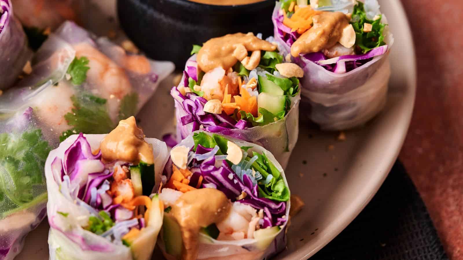Fresh spring rolls filled with shrimp, purple cabbage, carrots, greens, and topped with chopped peanuts and peanut sauce, served on a plate.