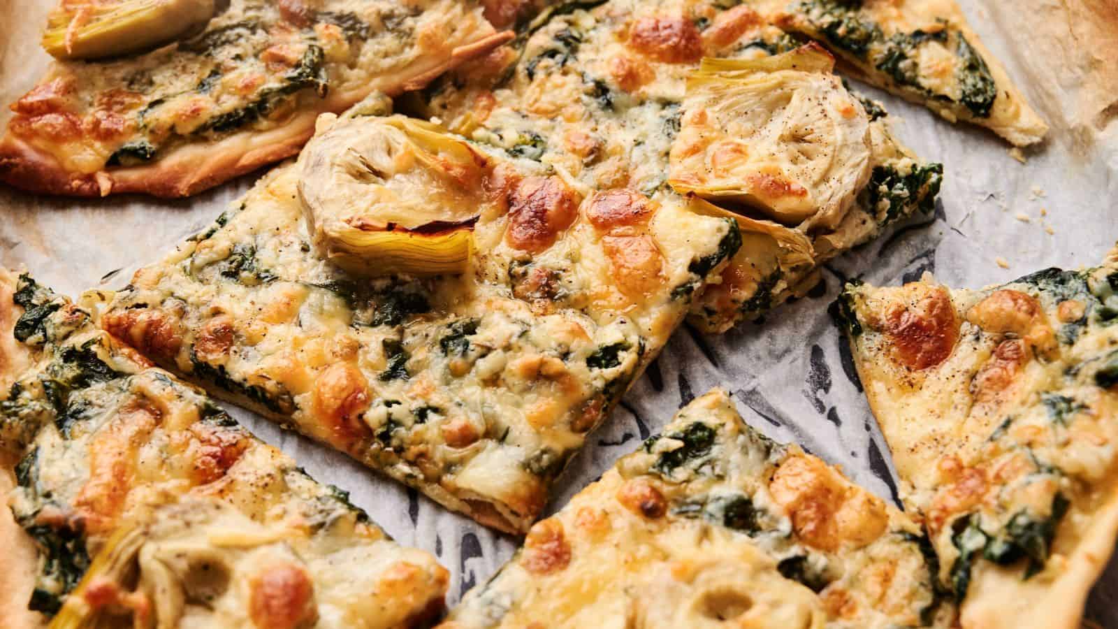 Rectangular slices of pizza topped with spinach, artichoke hearts, melted cheese, and a golden crust on parchment paper.