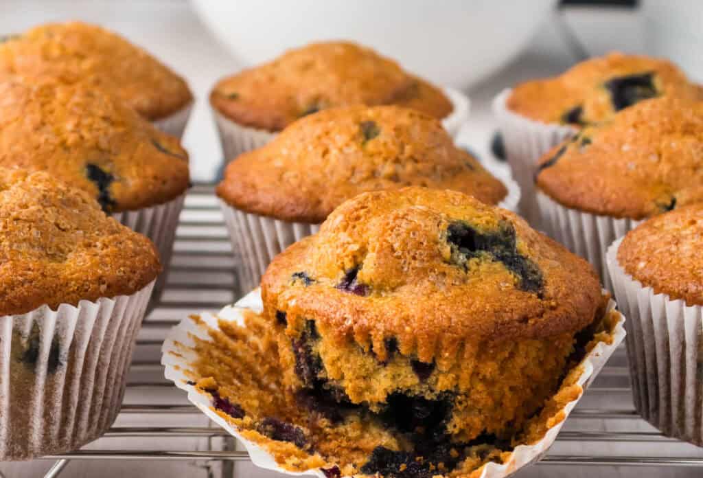A group of blueberry muffins, some unwrapped, sit on a metal cooling rack.