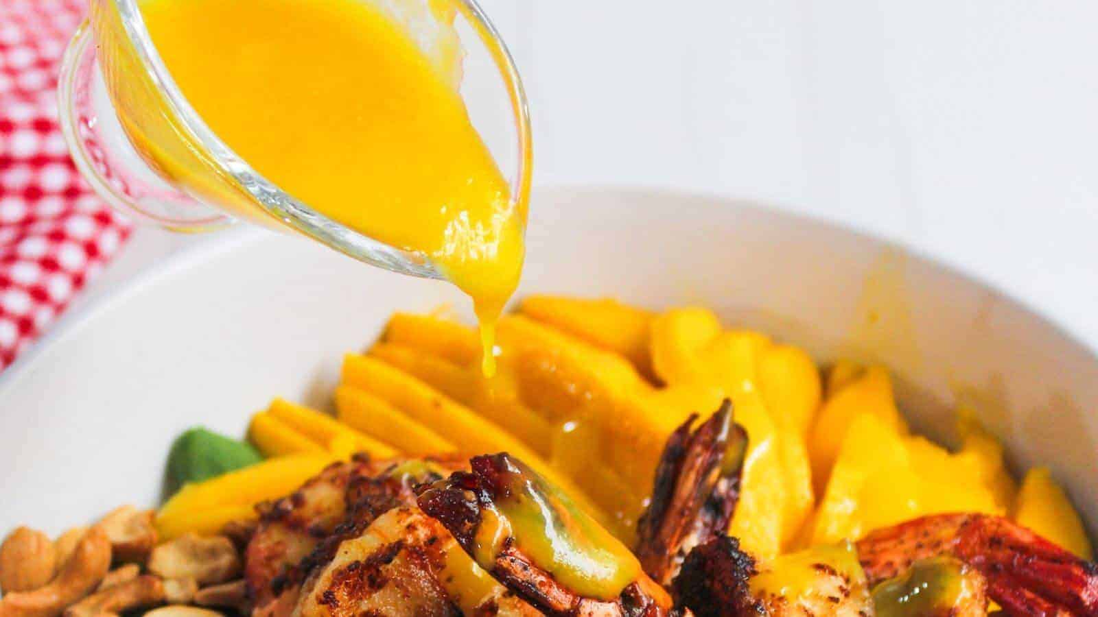 A glass container pours yellow sauce over grilled shrimp in a bowl with mango slices, nuts, and leafy greens.