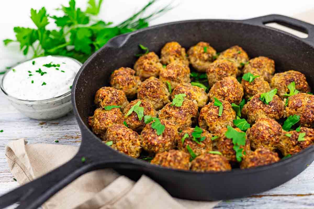 A cast iron skillet filled with cooked meatballs garnished with parsley sits on a napkin, with a bowl of white dipping sauce and fresh herbs in the background.