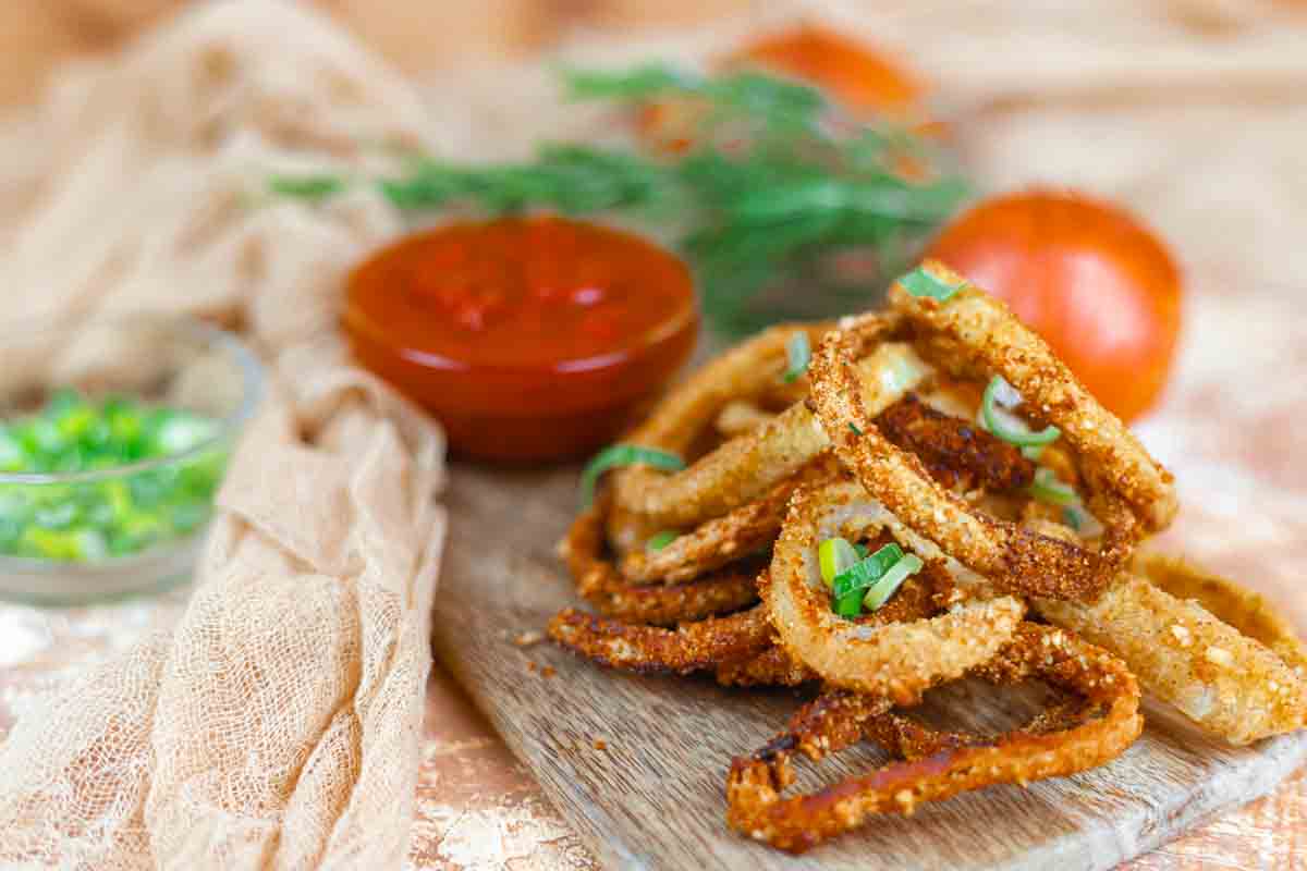 A pile of crispy fried onion rings garnished with green onions sits on a wooden board, with a bowl of red dipping sauce and a tomato in the background.
