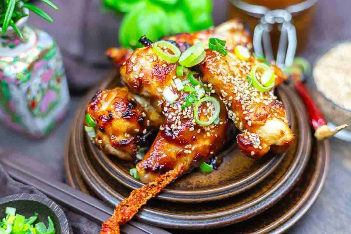 Oven baked BBQ wings garnished with sesame seeds and sliced green onions, served on a brown ceramic plate with decorative items in the background.