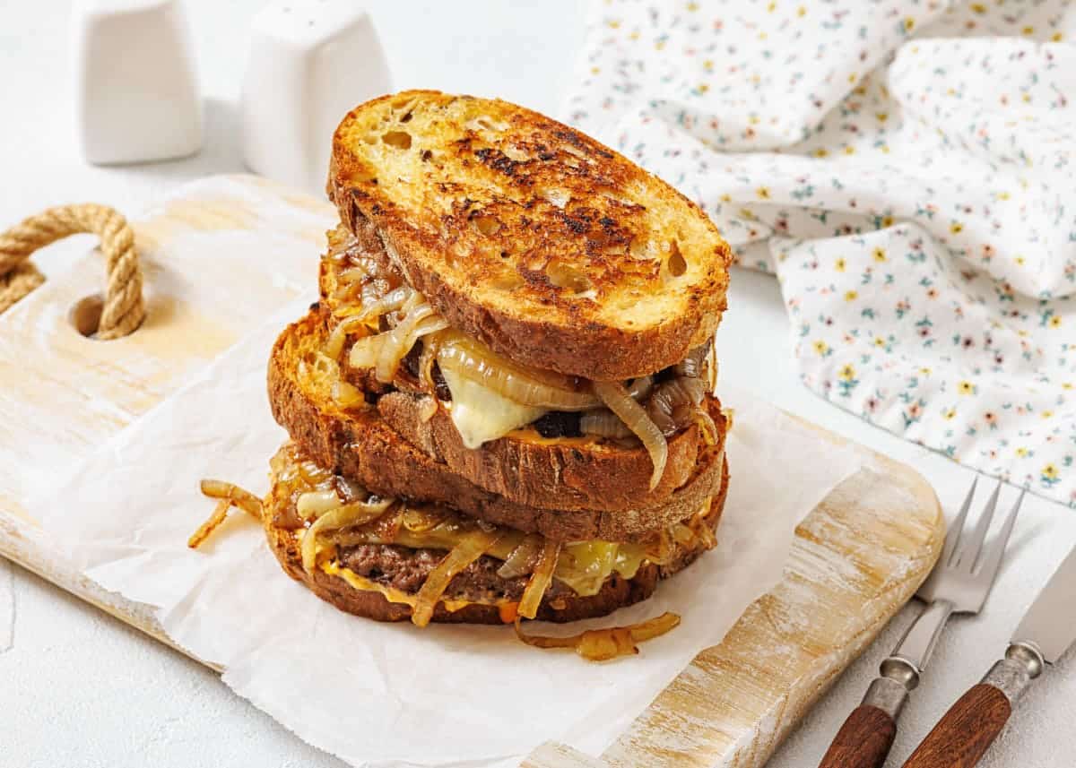 A stack of two patty melt sandwiches with caramelized onions and melted cheese on toasted bread, placed on parchment paper atop a wooden board.