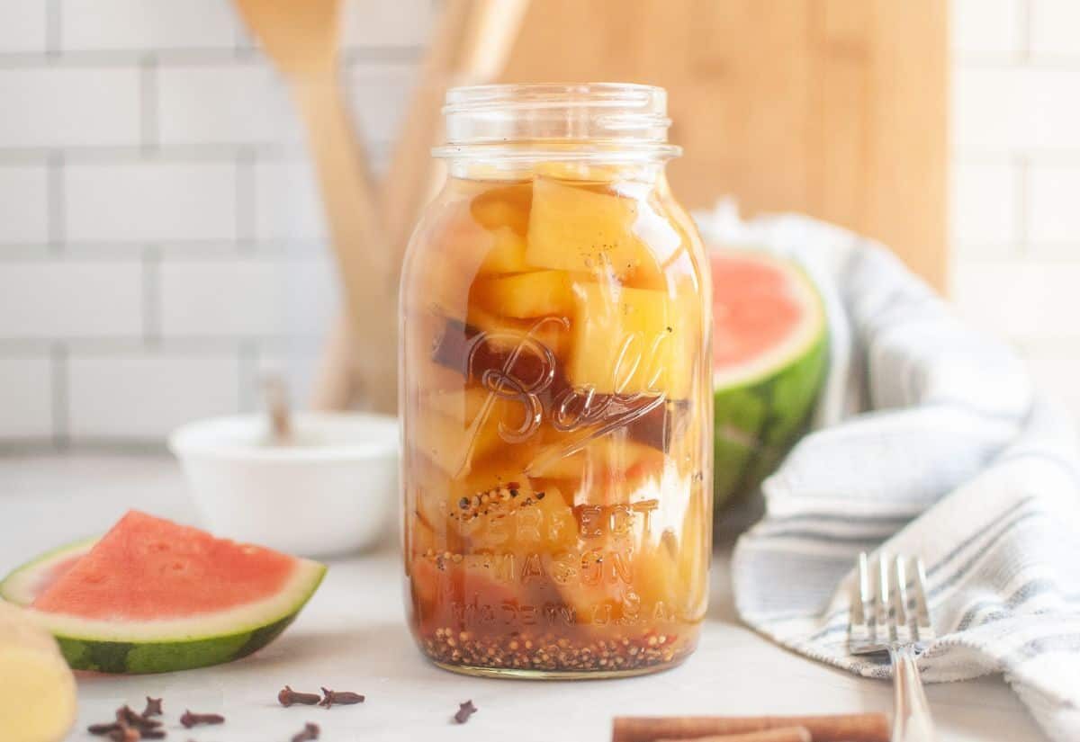 Jar of pickled watermelon rind and spices on a kitchen counter; nearby, a watermelon wedge and a striped towel enhance the fresh, rustic setting.