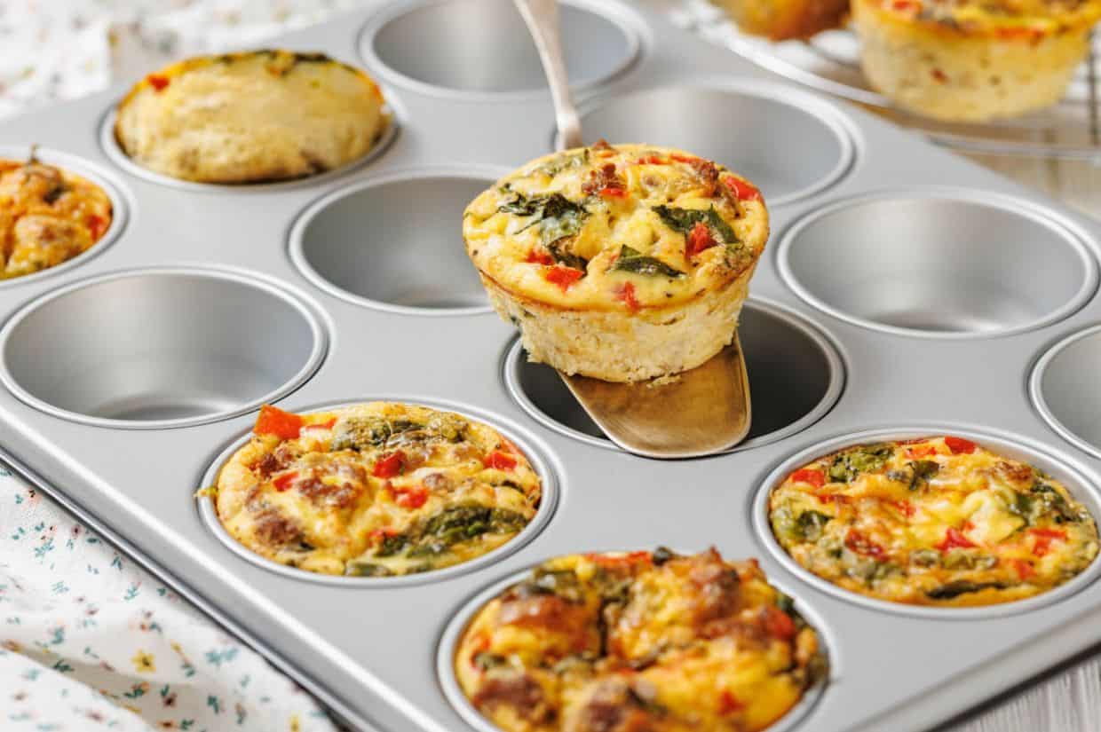 A metal muffin tin holds several breakfast baked egg muffins with vegetables; one is being lifted out with a spatula.