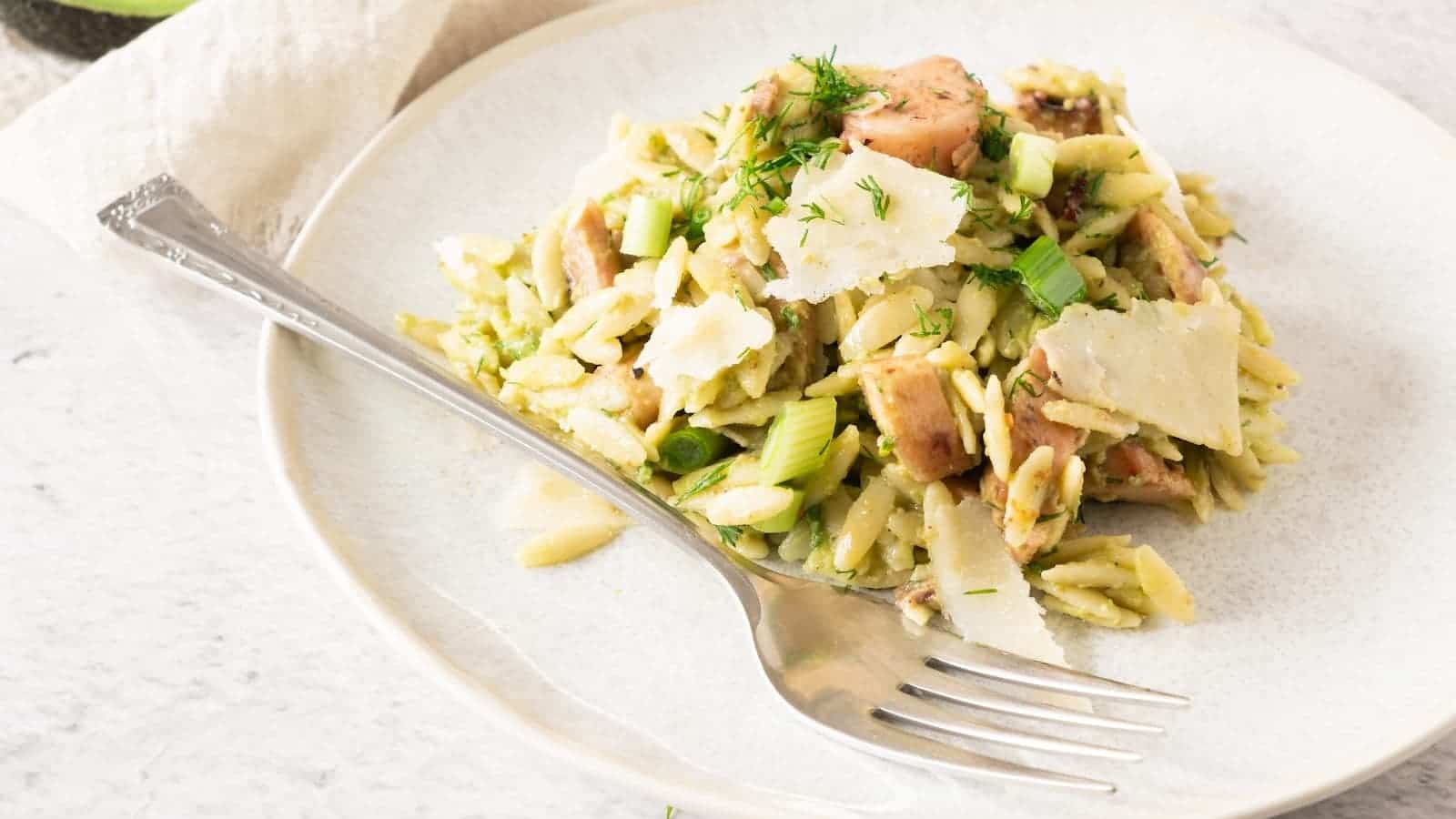A plate of orzo salad mixed with pieces of seafood, green vegetables, and topped with shaved parmesan cheese, served with a fork.