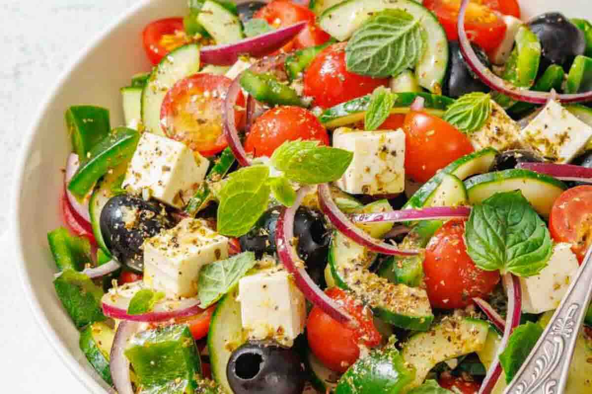 A bowl of salad with tomatoes, cucumbers, olives, red onion, feta cheese cubes, and fresh mint leaves, seasoned with herbs.