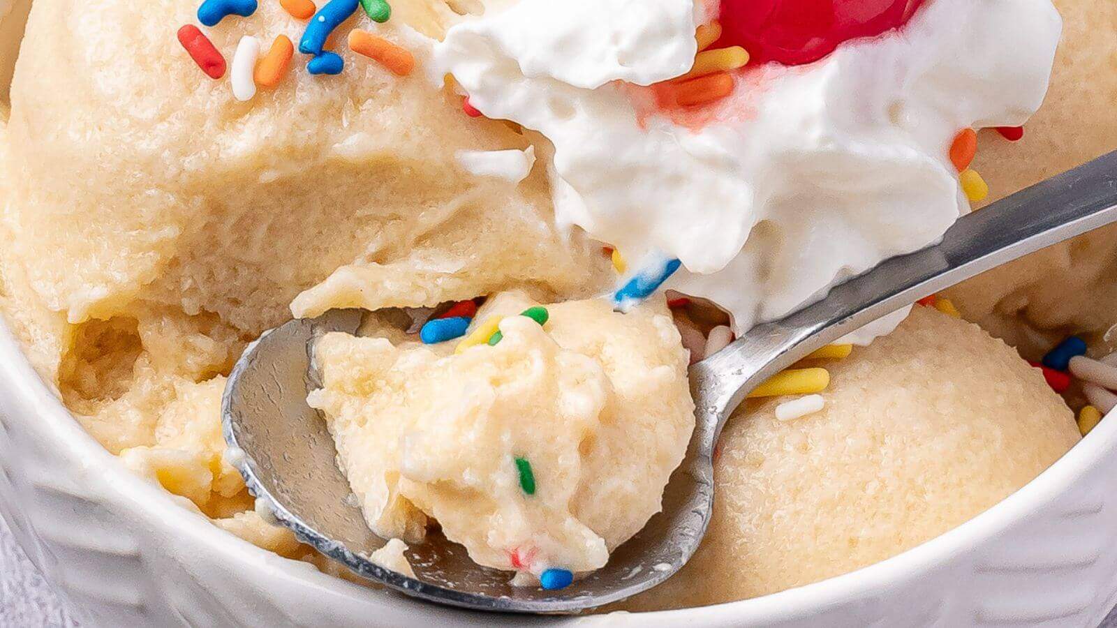 Close-up of a bowl of vanilla ice cream topped with whipped cream, a cherry, colorful sprinkles, and a spoon scooping a portion.