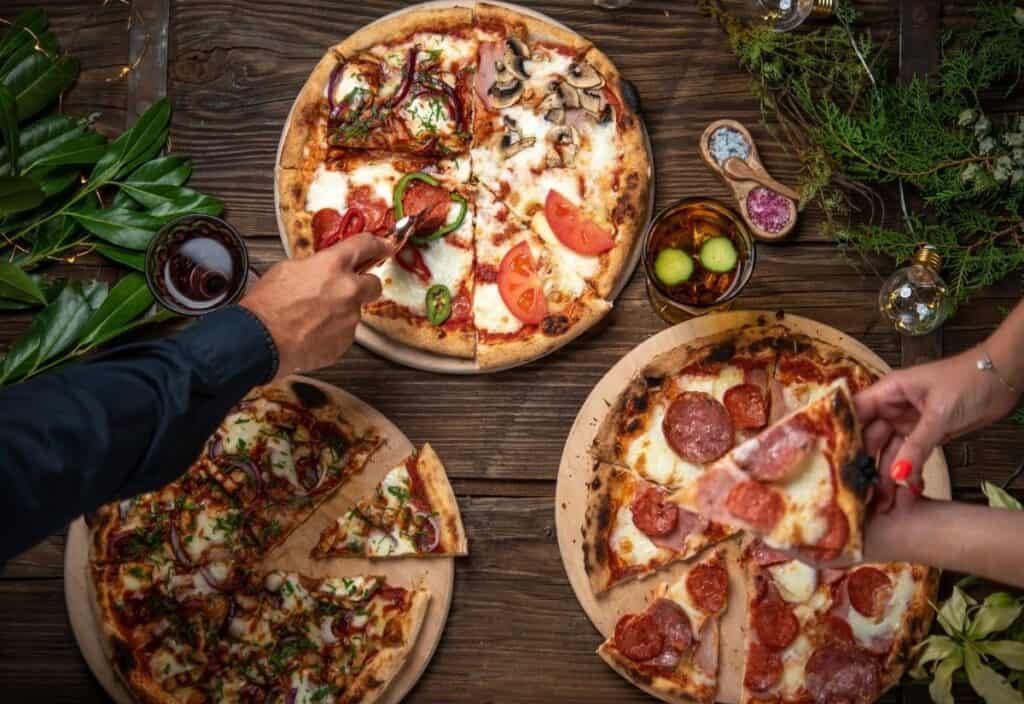 Three pizzas with various toppings on wooden boards, two hands taking slices, surrounded by drinks, olives, and greenery on a rustic wooden table&mdash;perfect for a feast made fresh from your backyard pizza oven.