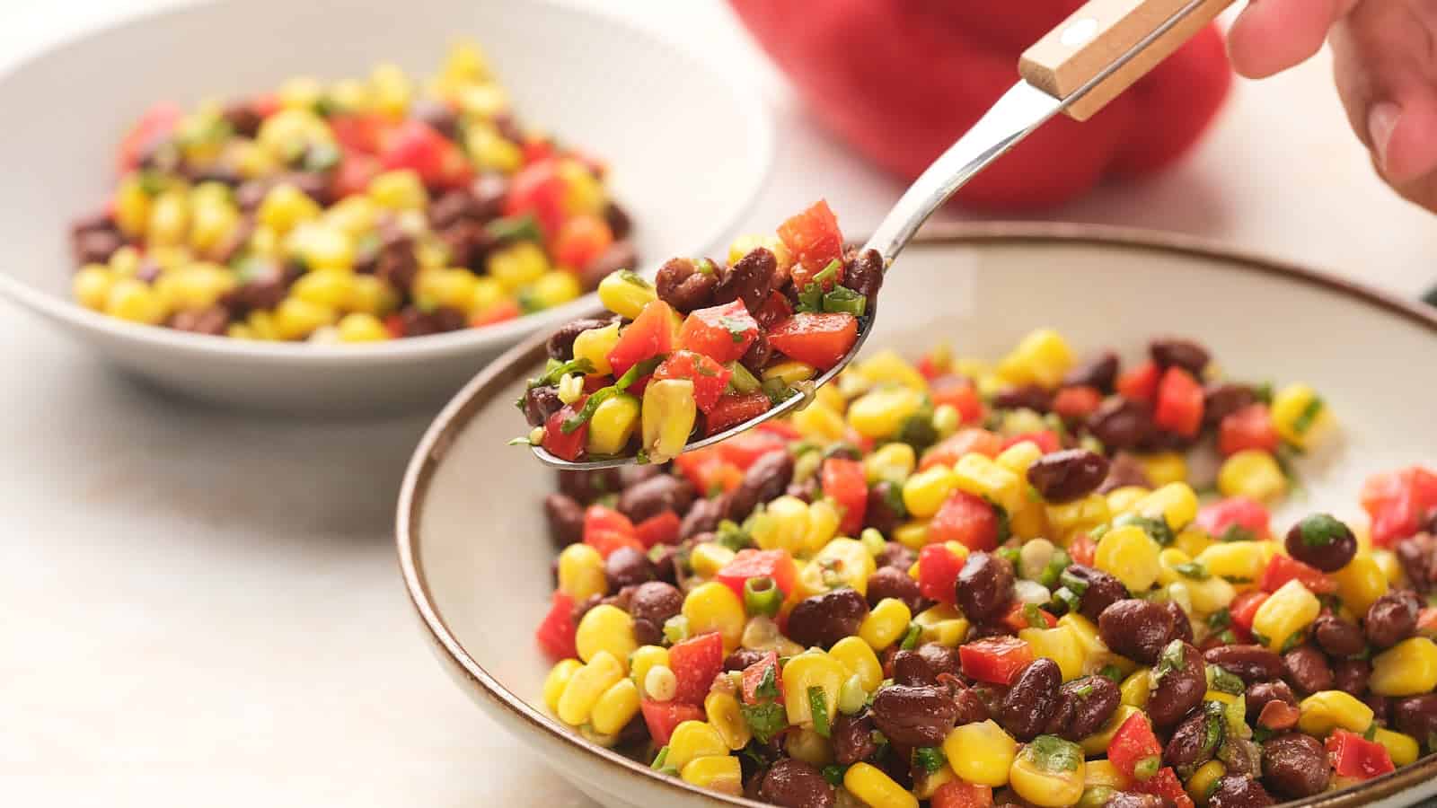 A bowl of black bean salad sits on a table with a spoon and a second small bowl in the background.