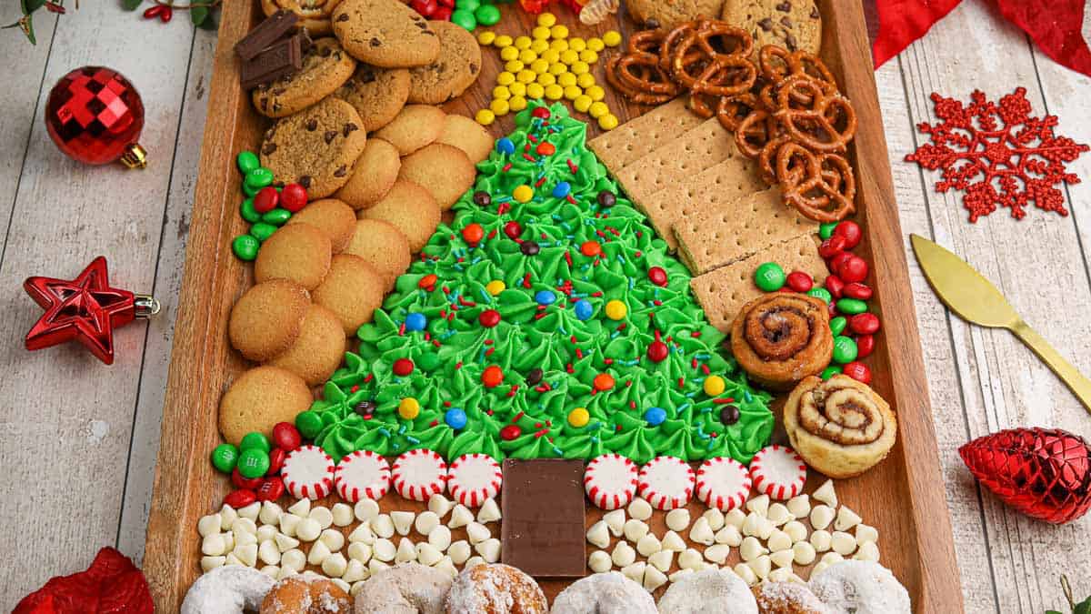 A festive Christmas-themed snack board with a tree made of green frosted cookies and an assortment of other treats, including cookies, pretzels, graham crackers, candy, and pastries, arranged around it.