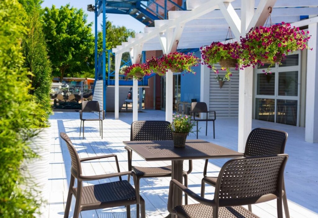 Outdoor patio area with brown tables and chairs on a white wooden deck, hanging flower baskets above, and trees in the background on a sunny day&mdash;perfect for enjoying one of the top restaurants in Charleston.