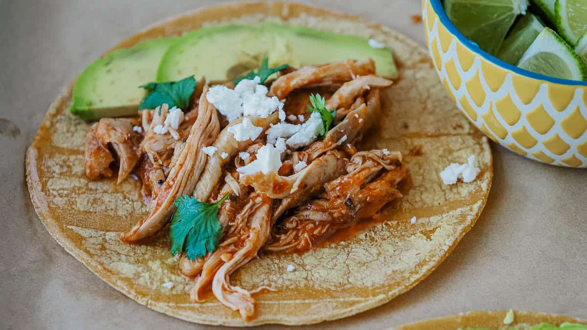 A corn tortilla topped with shredded chicken, avocado slices, cilantro, and crumbled cheese, next to a bowl of lime wedges.