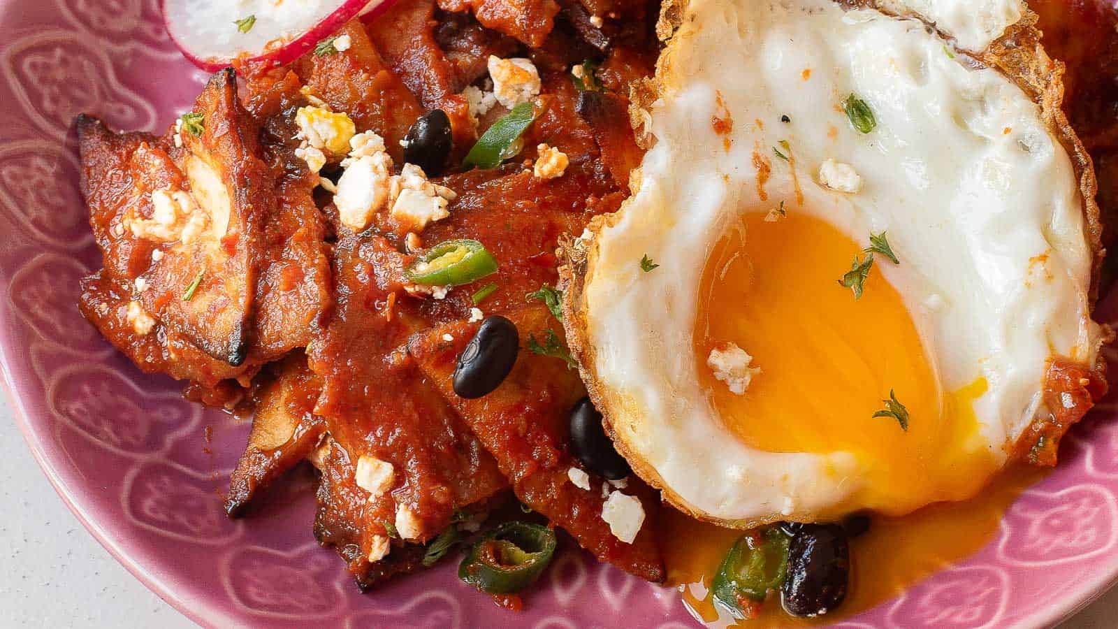 A close-up of a pink patterned dish featuring saucy grilled meat, fried eggs, black beans, green onions, and crumbled cheese—perfect for fans of a hearty Chilaquiles Rojos recipe.