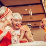 Three older women sit at a table, laughing and enjoying drinks and dessert together in a warmly lit indoor setting.