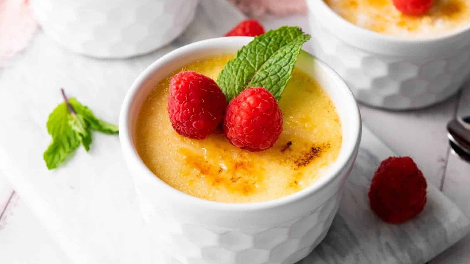 A ramekin of cr&egrave;me br&ucirc;l&eacute;e topped with two fresh raspberries and a mint leaf.