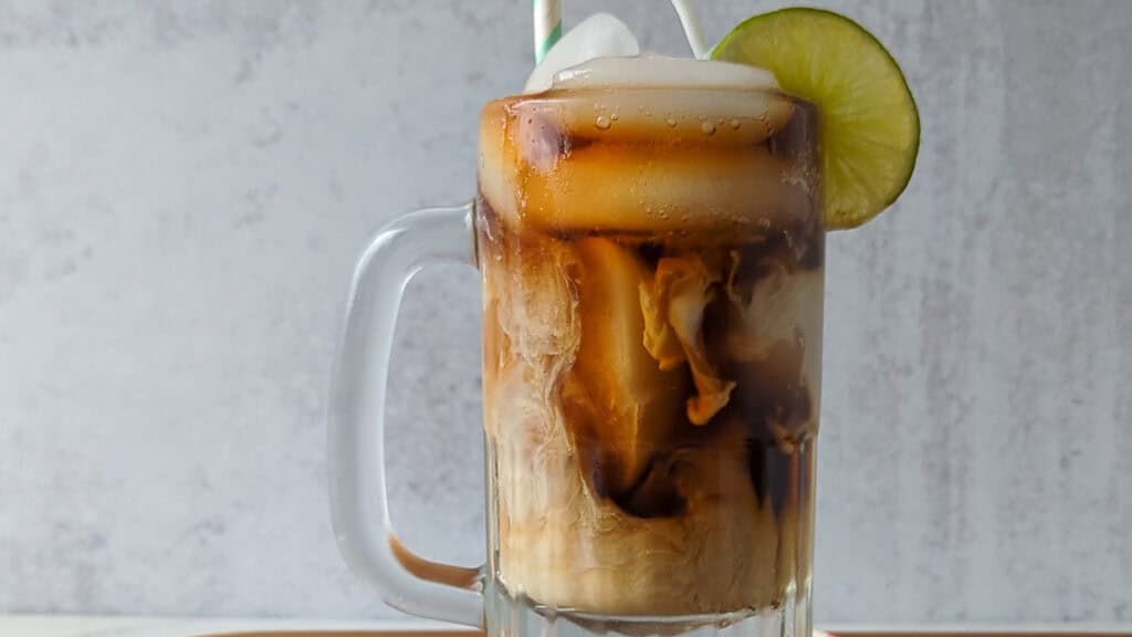 A glass mug filled with iced coffee, milk swirling inside, topped with ice, a lime slice on the rim, and a striped straw.