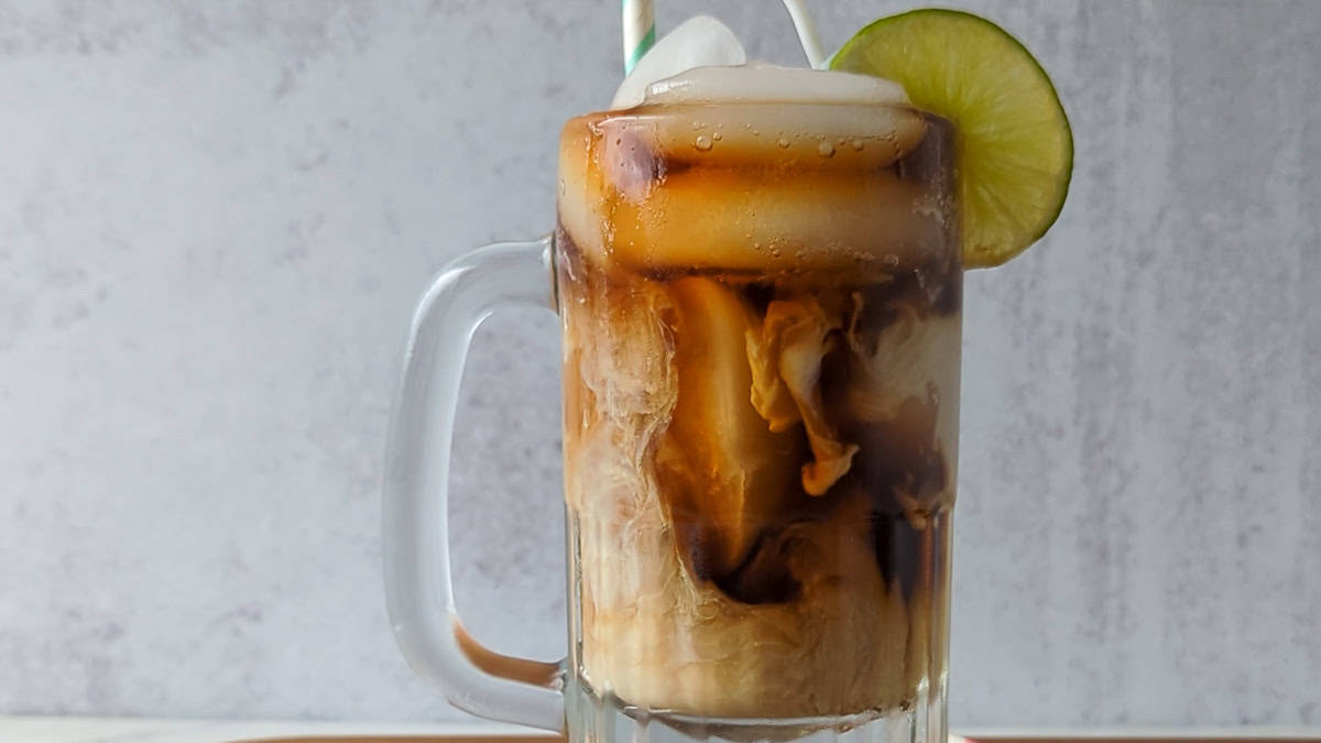 A glass mug filled with iced coffee, milk swirling inside, topped with ice, a lime slice on the rim, and a striped straw.
