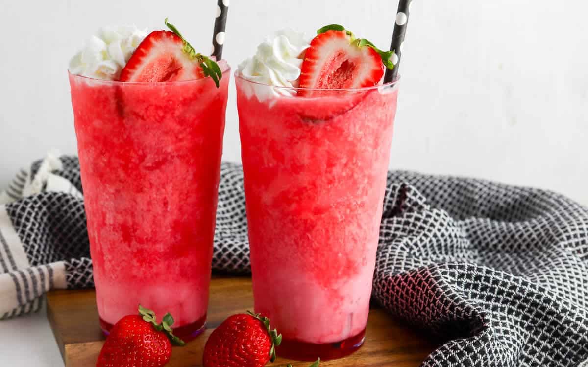 Two tall glasses filled with dirty strawberry soda, topped with whipped cream and sliced strawberries, each with a black and white polka dot straw. Fresh strawberries are placed beside the glasses.