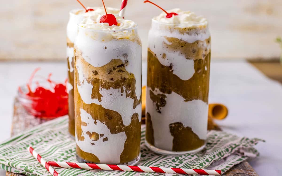 Three glasses of fluffy coke topped with whipped cream and a cherry on a patterned napkin, with red and white straws beside the glasses.