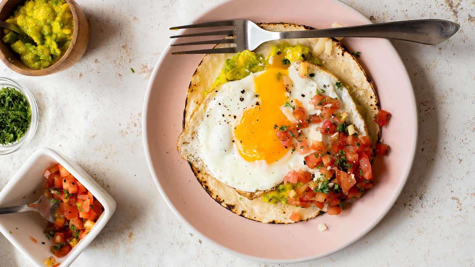 A plate featuring a delicious Fried Egg Taco on a tortilla, topped with creamy guacamole and fresh tomato salsa, with a fork and knife beside it.
