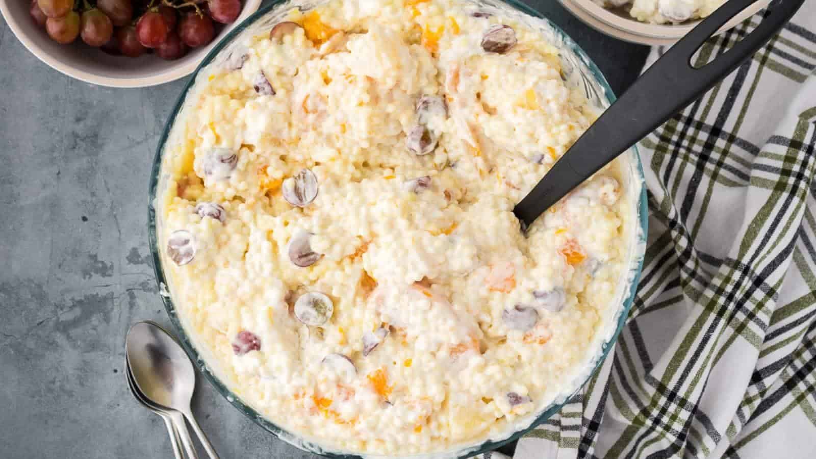 A bowl of creamy frog eye salad sits on a table next to a stripe cloth napkin and spoons.