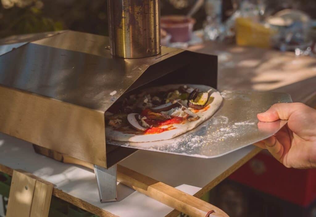 A hand uses a metal peel to place a homemade vegetable pizza into a small backyard pizza oven.