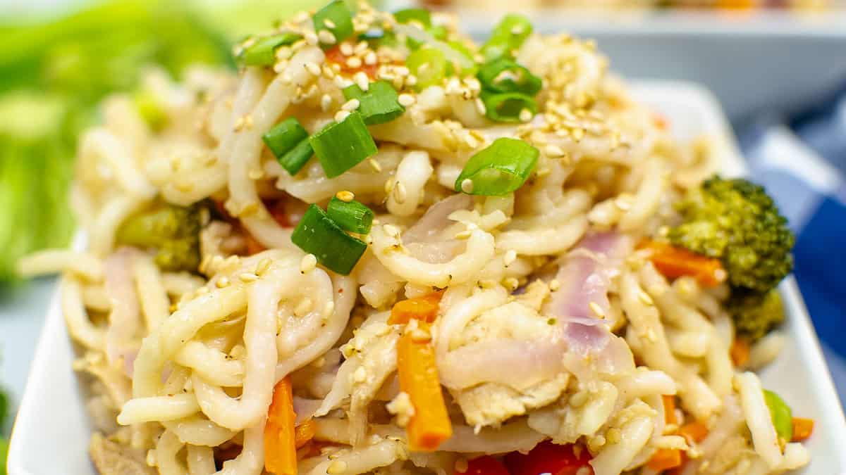 Close-up of a bowl of stir-fried noodles topped with chopped green onions, sesame seeds, chicken, carrots, broccoli, and red onion.