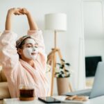 On international self care day, a person wearing a face mask and bathrobe stretches with eyes closed while sitting in front of a laptop, with tea and snacks on the table.