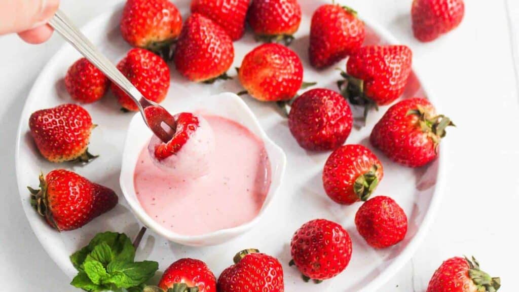 A plate of fresh strawberries surrounds a small bowl of pink strawberry dip, with a hand dipping one strawberry into the sauce using a fork.