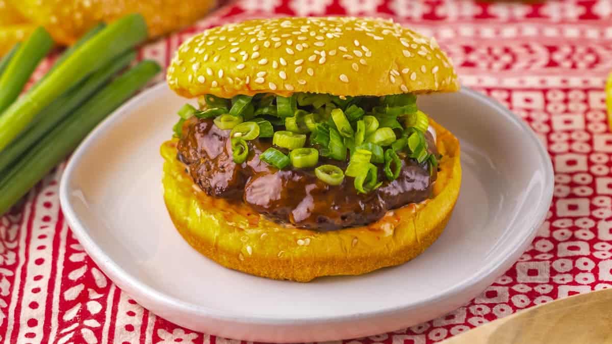 A burger with a sesame seed bun, topped with a beef patty, sauce, and a generous amount of chopped green onions, served on a white plate against a patterned red and white tablecloth background.