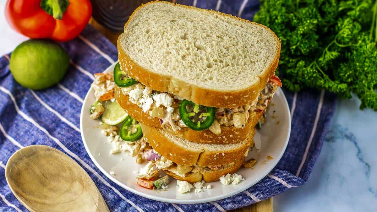 A plate with stacked sandwiches filled with vegetables, cheese, and jalapeños on a striped cloth, surrounded by a tomato, lime, parsley, and a wooden spoon.