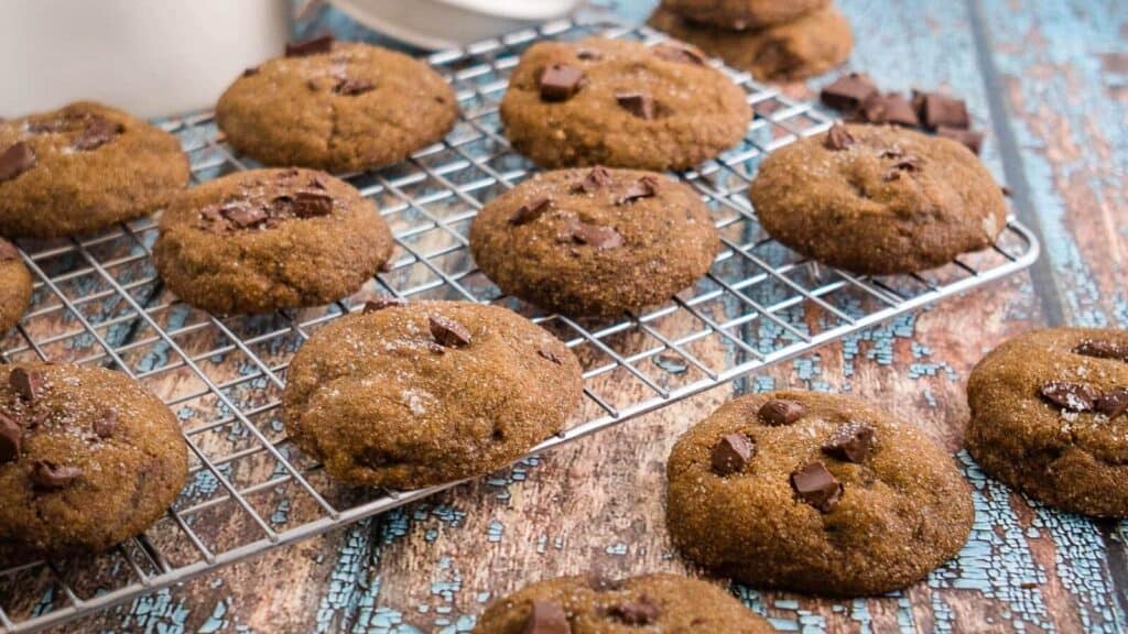 A cooling rack with chocolate chunk cookies on top, and several cookies placed on a rustic blue wooden surface.