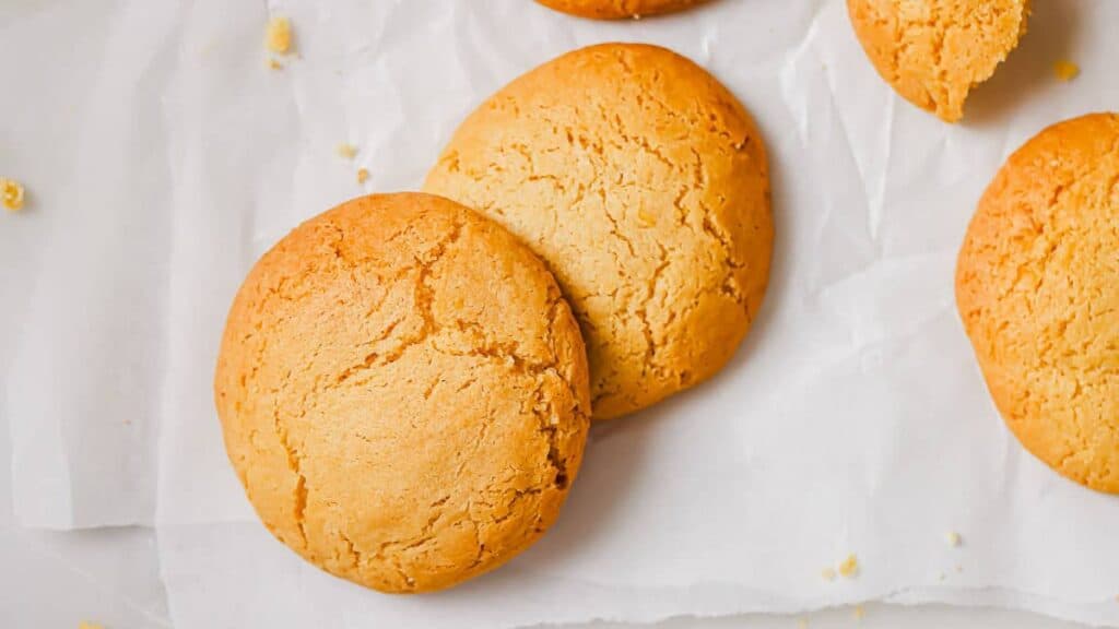 Two golden brown cookies resting on a sheet of white parchment paper, with a partial cookie and crumbs visible nearby.