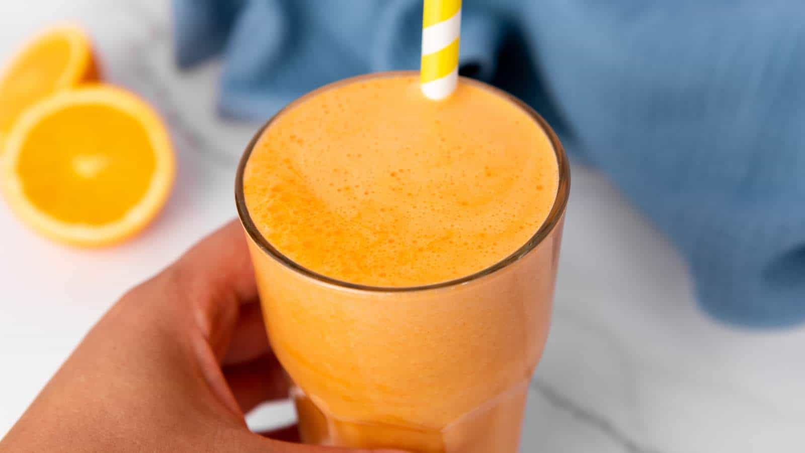 A hand holds a glass of orange smoothie with a yellow and white striped straw.