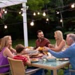 Six people sit around an outdoor dining table in a garden, sharing a meal under string lights in the evening.
