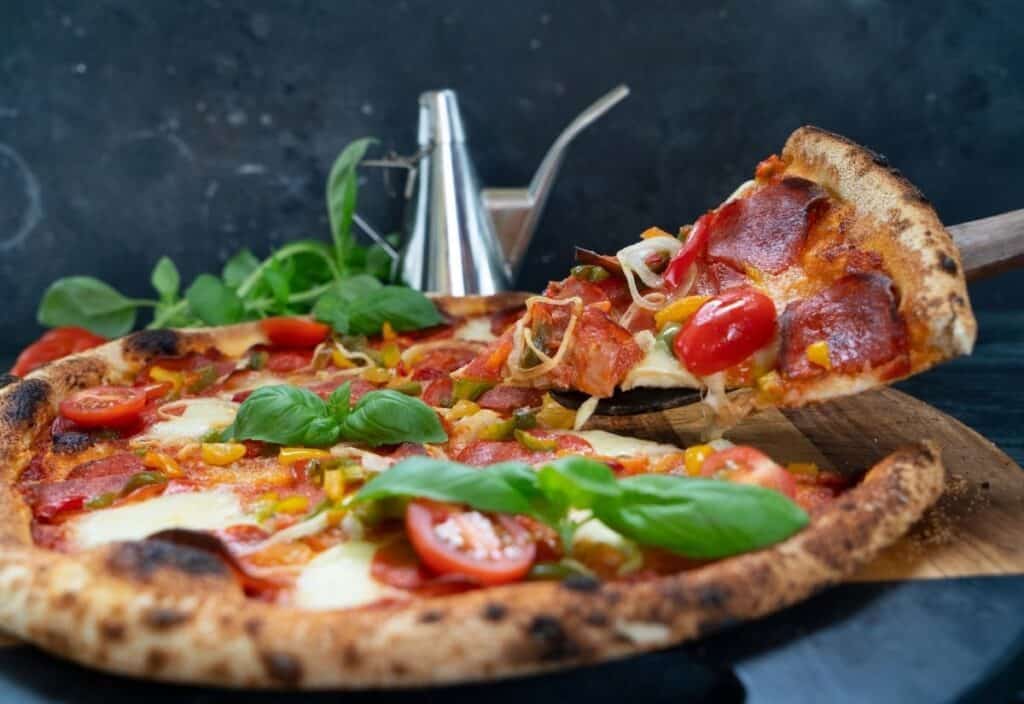A wood-fired pizza with pepperoni, tomatoes, cheese, and basil sits on a wooden board. Fresh from the backyard pizza oven, a metal spatula lifts one slice while an oil dispenser is in the background.