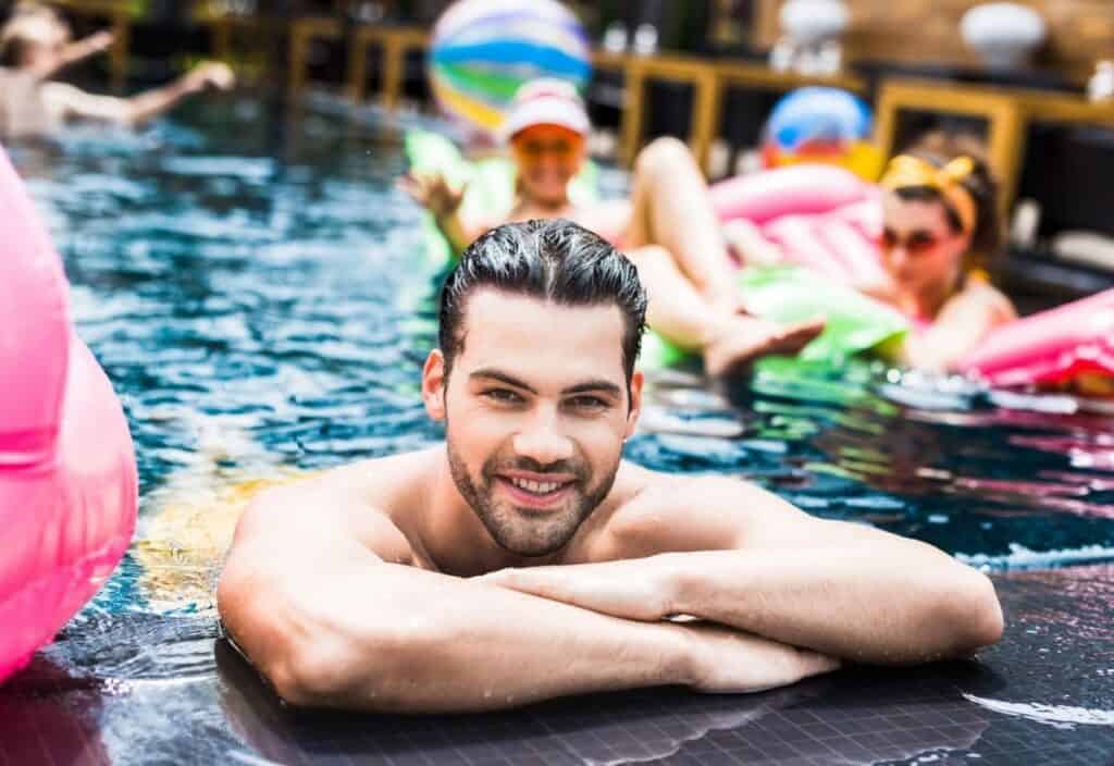 Man with slicked-back hair smiles at the edge of a swimming pool, with people relaxing on inflatables in the background.