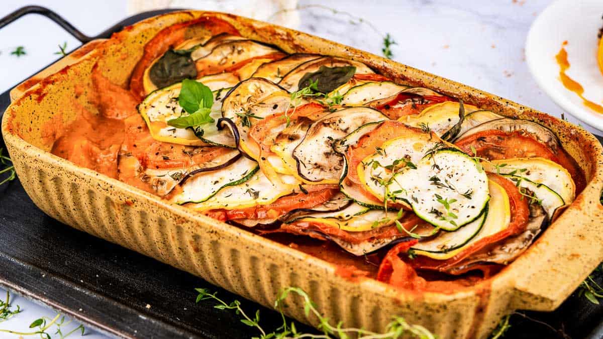 A rectangular baking dish filled with neatly layered slices of zucchini, tomato, and eggplant, garnished with herbs.