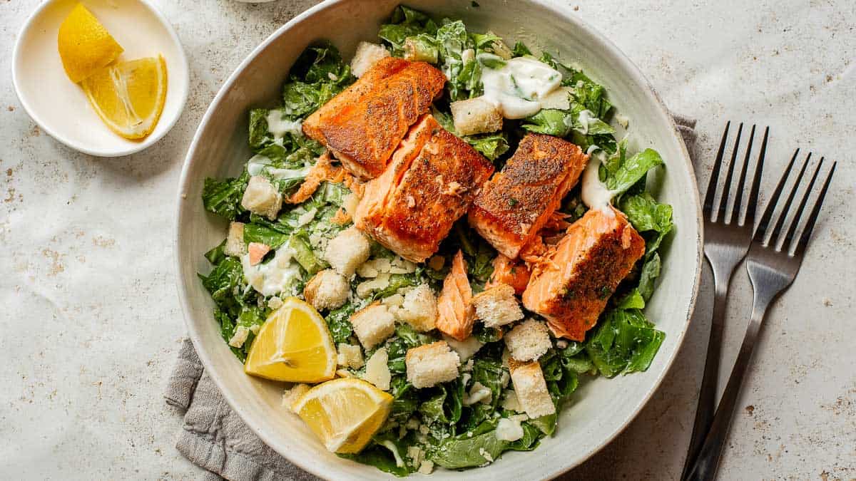 A bowl of Caesar salad topped with grilled salmon fillets, croutons, and lemon wedges, with Caesar dressing drizzled on top. Two forks and extra lemon are on the side.