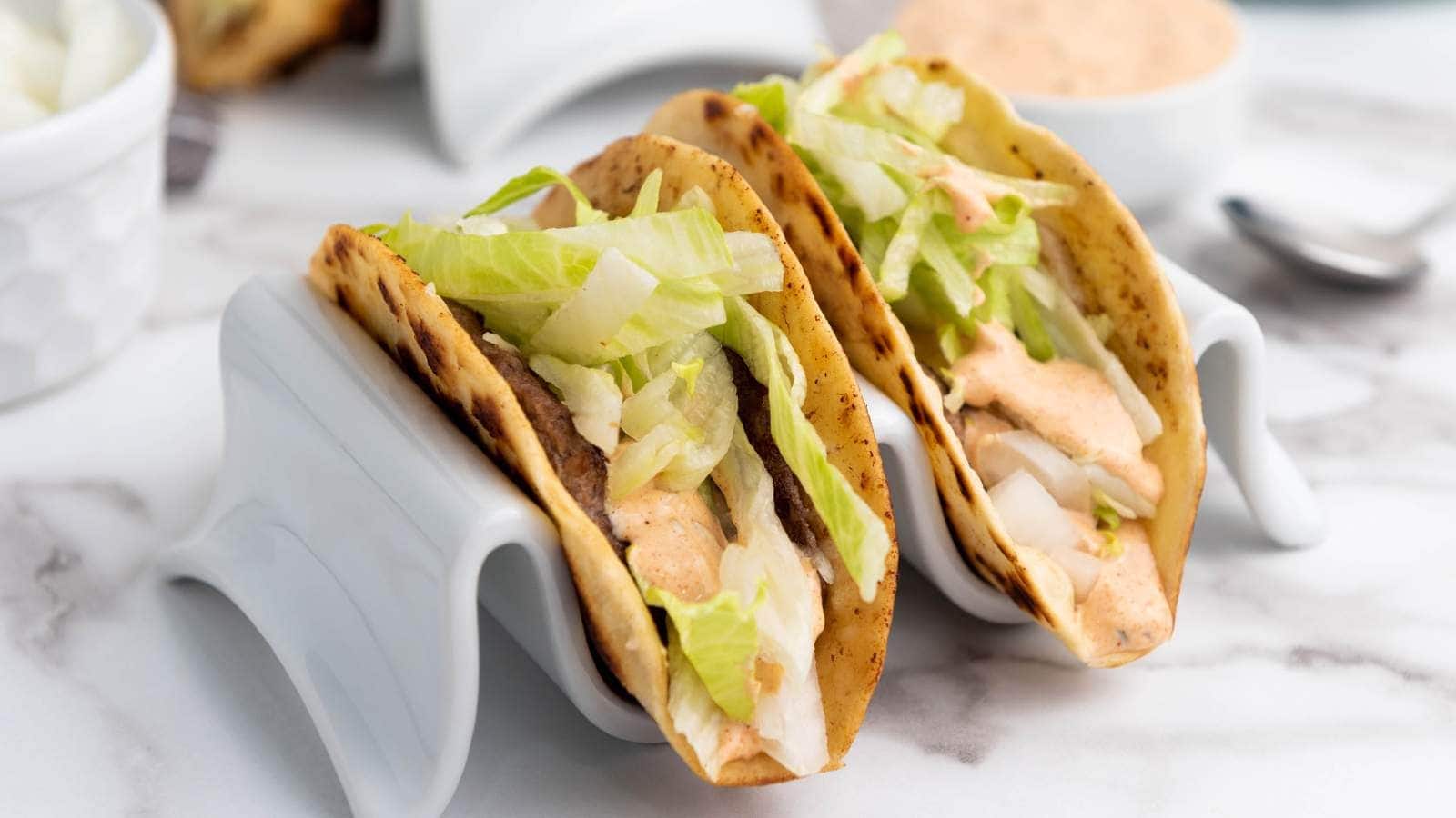 Two smash burger taco in taco holders topped with shredded lettuce, diced onions, and a creamy sauce.