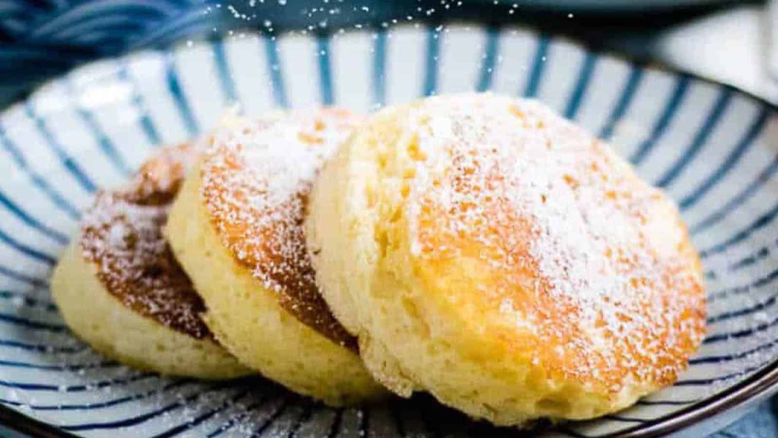 Four round Japanese souffle pancakes dusted with powdered sugar are arranged on a striped plate, with additional pastries on a patterned plate nearby. Persimmons are visible in the upper left corner.