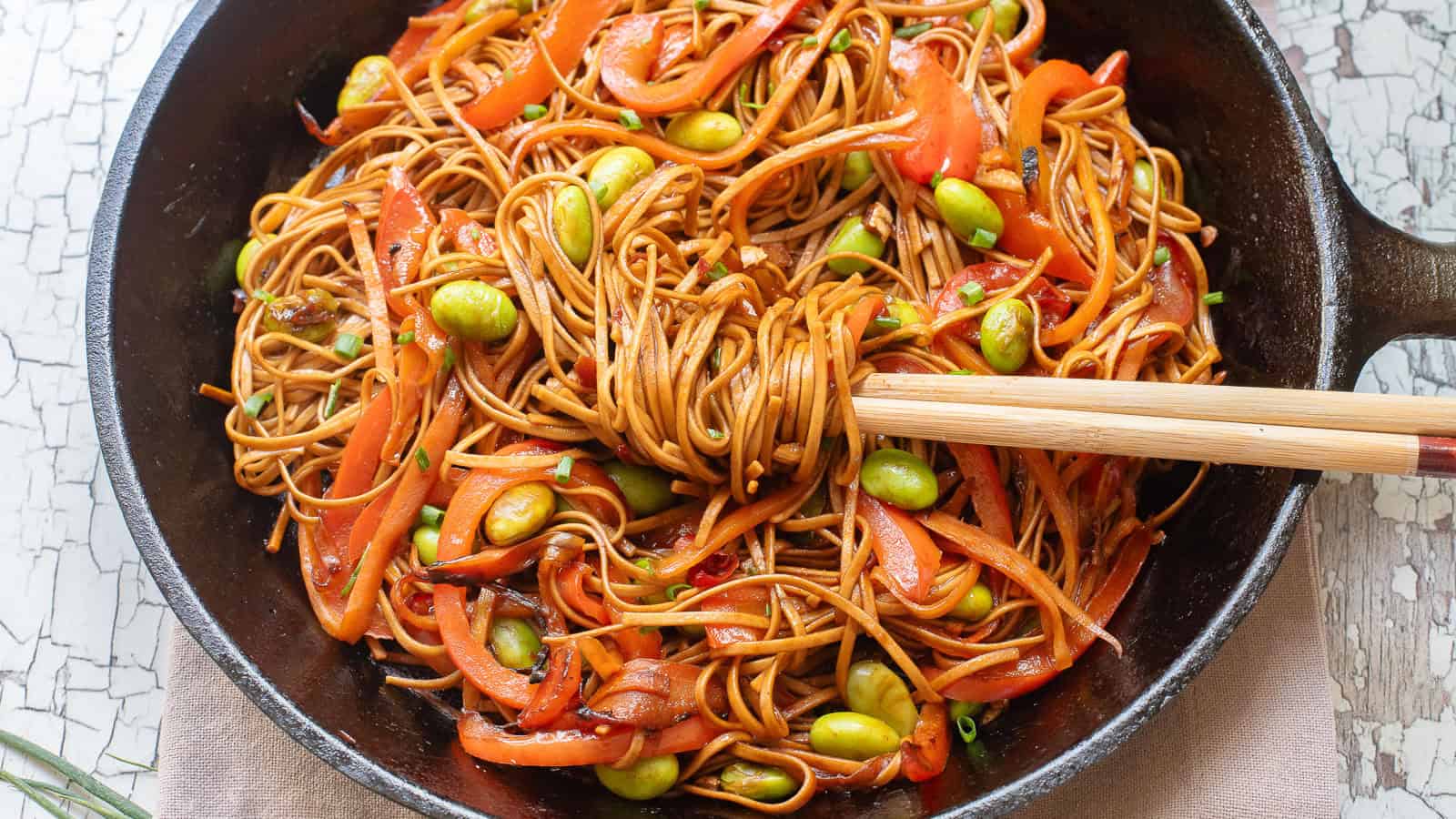 A purple bowl filled with noodles tossed with sliced red bell peppers, edamame, and green onions on a rustic white wooden surface. A few carrot slices and green onion leaves are partially visible on the side.